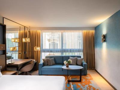 Chambre d'hôtel moderne avec lit double, canapé, bureau et grandes fenêtres avec rideaux.