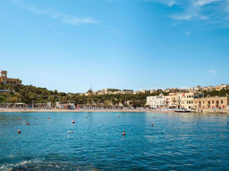 Ville côtière avec eau bleue, bâtiments au bord de l'eau et collines boisées en arrière-plan.
