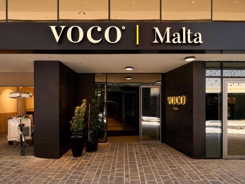 Entrée d'hôtel moderne avec cadre sombre et panneau Voco Malta au-dessus.
