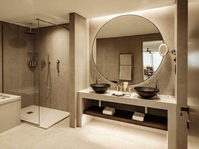 Nowoczesna łazienka z dużym okrągłym lustrem, podwójną umywalką i otwartą kabiną prysznicową.