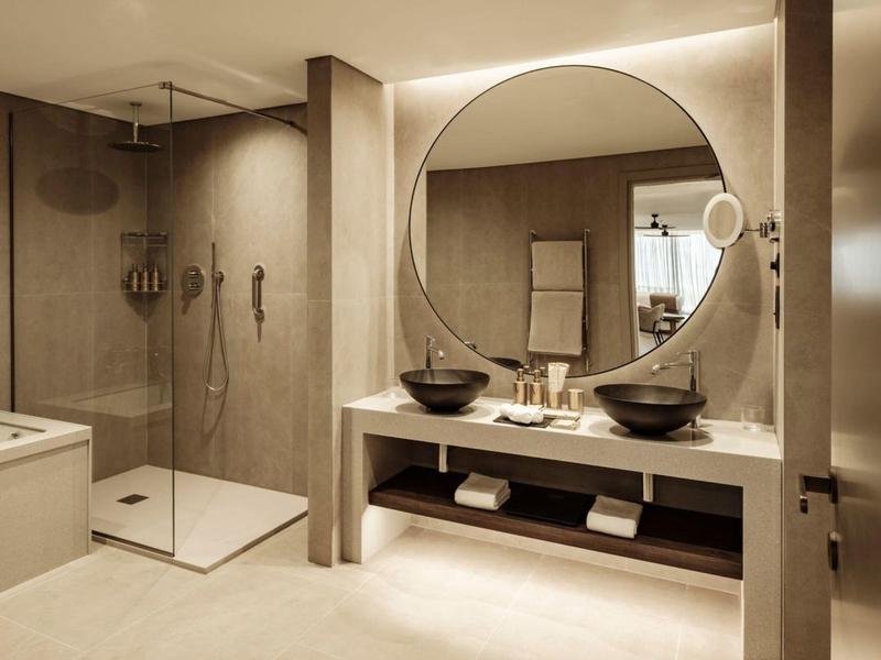 Nowoczesna łazienka z dużym okrągłym lustrem, podwójnymi umywalkami i prysznicem walk-in.