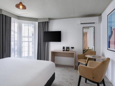 Camera d'albergo moderna con letto grande, TV, scrivania e due poltrone alla luce del giorno.