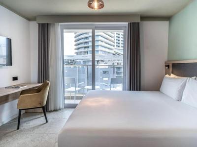 Camera d'hotel moderna con letto grande, scrivania, sedia e balcone con vista su altri edifici.