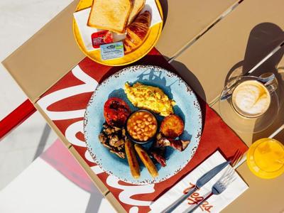 Piatto con colazione composta da uova strapazzate, fagioli al forno, pancetta e toast su un tavolo con caffè e succo.