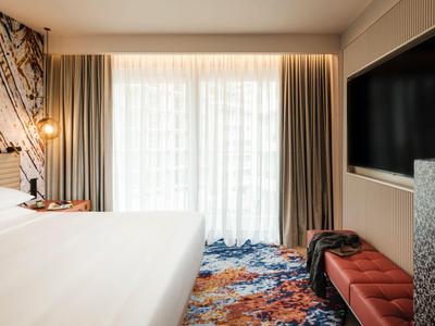 Hotelzimmer mit großem Bett, Fenster mit Vorhängen und buntem Teppichboden.