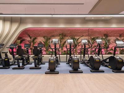 Modern fitnesscentrum met meerdere fitnessapparaten en palmbomen in een hotelinterieur.