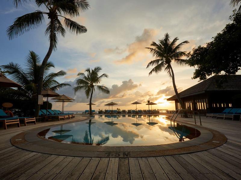 Runder Pool mit Holzdeck, Palmen und Liegestühlen bei Sonnenuntergang am Meer.