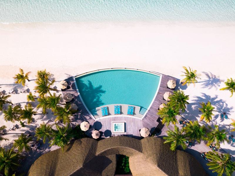 Blick von oben auf einen tropischen Pool mit Liegestühlen und Palmen am Sandstrand.