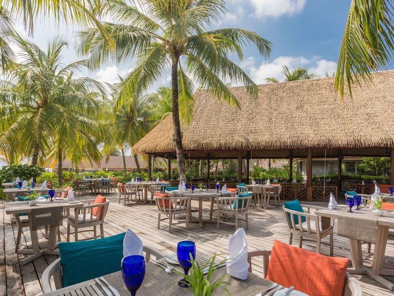 Freiluftrestaurant mit Strohdach und bunten Stühlen unter Palmen bei sonnigem Wetter.