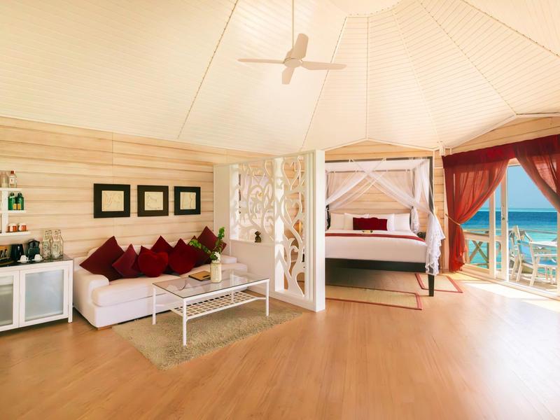 Helles Ferienzimmer mit Bett, Sofa, Ventilator und Blick auf das Meer.