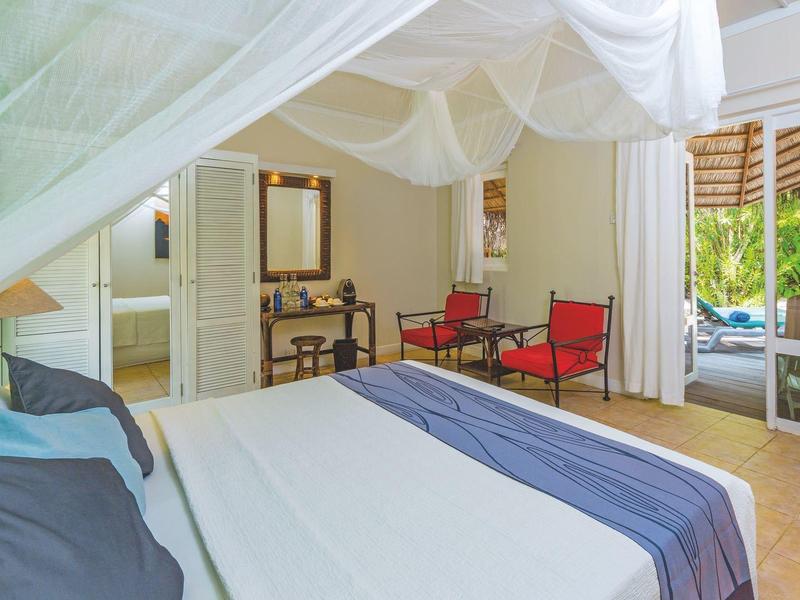 Hell eingerichtetes Schlafzimmer mit großem Bett, Sitzbereich und Blick auf einen Garten.