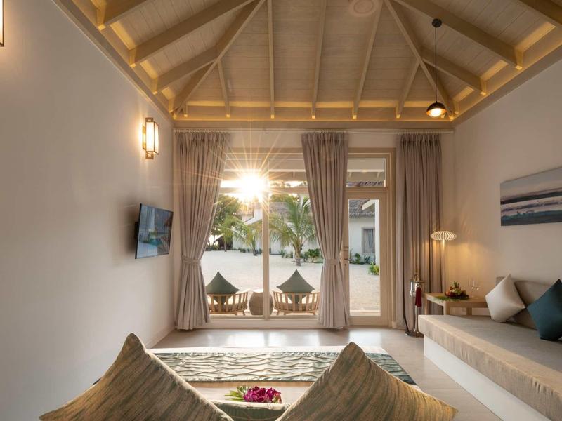 Gemütliches Wohnzimmer mit Holzdecke, großem Fenster, Sofa, Kissen und Blick auf grüne Außenanlage.