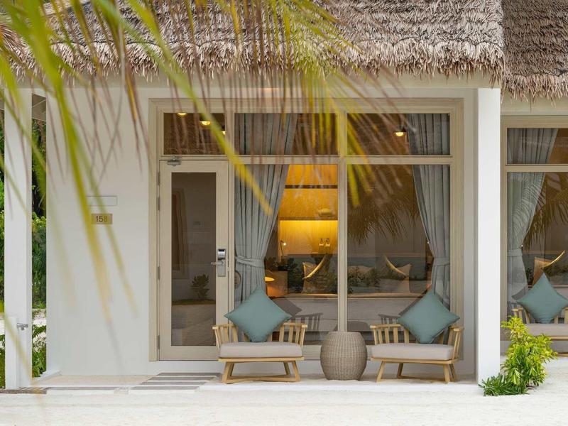 Terrassenbereich mit zwei Holzsesseln, blauen Kissen, Vase und Lampenlicht hinter großen Fenstern.