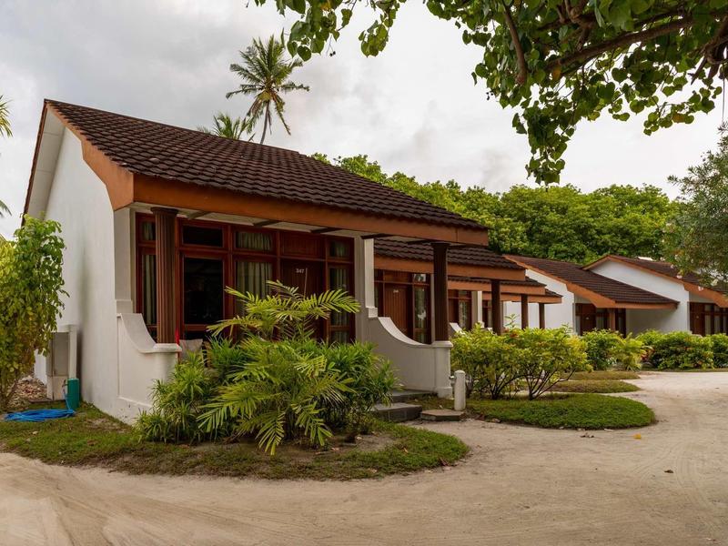 Weiße Bungalows mit braunen Dächern und Holzfenstern, umgeben von grünen Pflanzen und Sandweg.