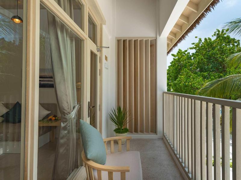 Balkon mit Holzmöbeln und weißen Handläufen, grüne Pflanzen und Bäume im Hintergrund.