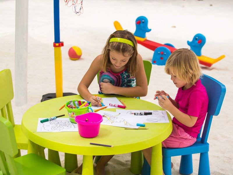 Zwei Kinder sitzen an einem runden grünen Tisch und malen mit Buntstiften auf Papier.