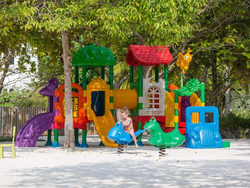 Bunte Klettergerüst und Rutschen auf einem Spielplatz mit Kindern und Bäumen im Hintergrund.
