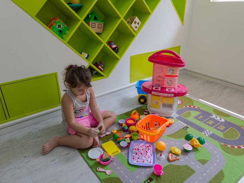 Ein Mädchen sitzt auf einer bunten Spielmatte und spielt mit Spielzeug in einem hellen Raum mit grünem Regal.