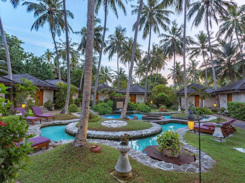 Tropische Gartenanlage mit Pool, Palmen und Gebäuden im Hintergrund bei klarem Himmel.