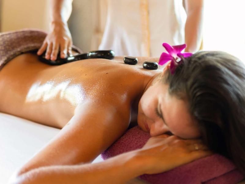 Frau liegt entspannt auf Massagebank, heiße Steine liegen auf ihrem Rücken, Blume im Haar.