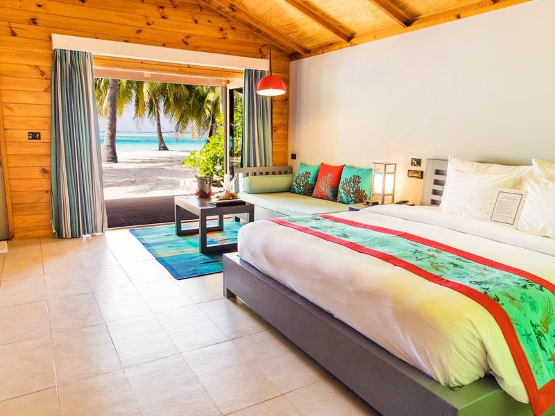 Helle Hotelzimmer mit großem Bett, bunten Kissen und Meerblick durch offene Tür.