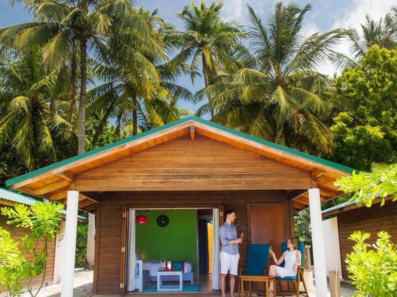 Holzhütte mit Überdachung und zwei Personen davor, umgeben von Palmen bei sonnigem Himmel.