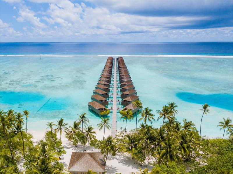 Überwässerte Bungalows auf türkisfarbenem Wasser mit Palmen und Sandstrand im Vordergrund.