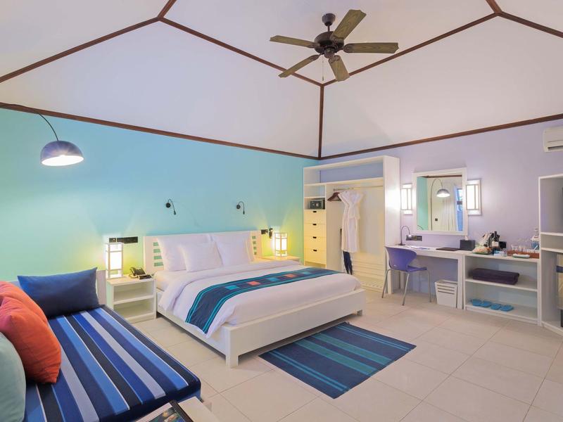 Helles Hotelzimmer mit großem Bett, blauer Wand, Sofa und Deckenventilator.