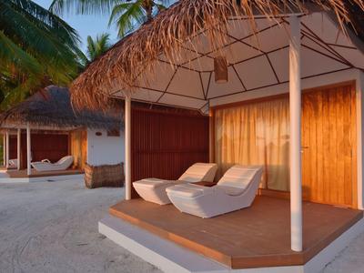 Strandbungalows mit Strohdächern, Holzwänden und weißen Lounge-Sesseln auf Sand bei Abendlicht.