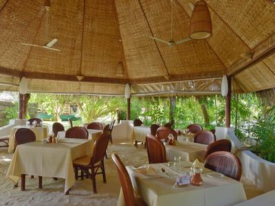 Offenes Restaurant mit Strohdach, gedeckten Tischen und Holzstühlen in grünem Tropengarten.