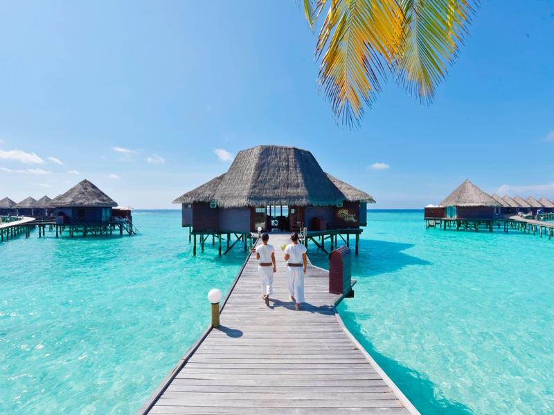 Ein Steg führt zu Überwasser-Bungalows über klarem, türkisfarbenem Wasser unter blauem Himmel.