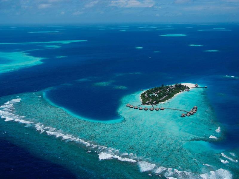 Kleine Insel mit grüner Vegetation, umgeben von türkisblauem Wasser und Korallenriffen im Ozean.