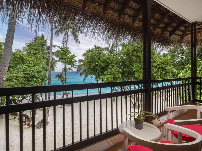 Balkon mit weißen Stühlen, Tisch und Pflanzen, Blick auf Sand, Bäume und blaues Meer.