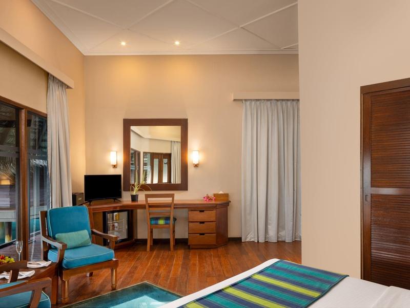 Hotelzimmer mit Holzboden, blauer Sitzgruppe, Schreibtisch, großem Spiegel und Kleiderschrank in warmen Farben.