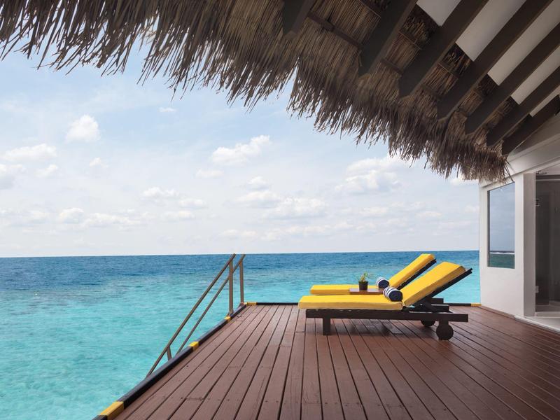 Terrasse mit Holzboden und gelben Liegestühlen über klarem blauem Ozean unter bewölktem Himmel.