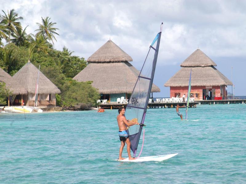 Person surft auf gleitendem Brett im türkisfarbenen Meer vor Insel mit Palmen und strohgedeckten Hütten.