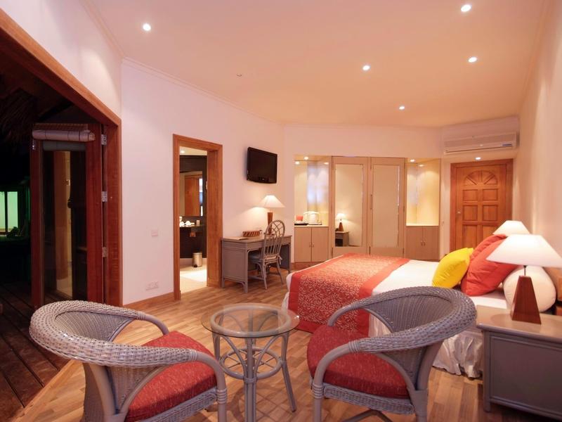 Wohnzimmer mit Holzboden, zwei Korbsesseln, Glas-Couchtisch, rotem Sofa und heller Wandbeleuchtung.