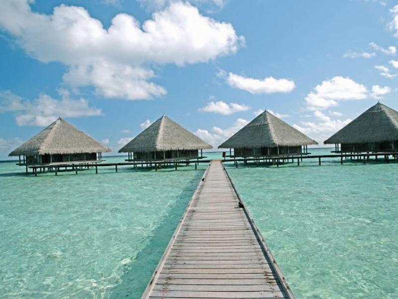 Vier über dem klaren, türkisfarbenen Wasser gebaute Bungalows mit einem Steg unter blauem Himmel.