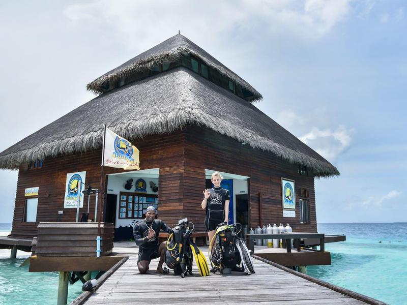 Holzhaus mit Strohdach auf Steg, Tauchausrüstung davor, umgeben von blauem Meer und Himmel.