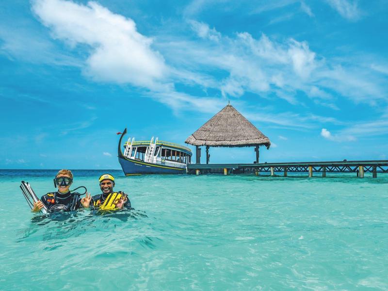 Zwei schnorchelnde Personen im klaren, türkisfarbenen Wasser vor einem Holzsteg mit Hütte und Boot, blauer Himmel.
