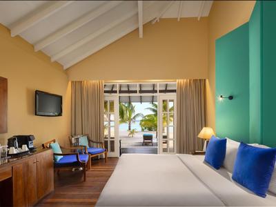 Helles Hotelzimmer mit großem Bett, blauen Kissen, Holzmöbeln und Blick auf Terrasse und Meer.