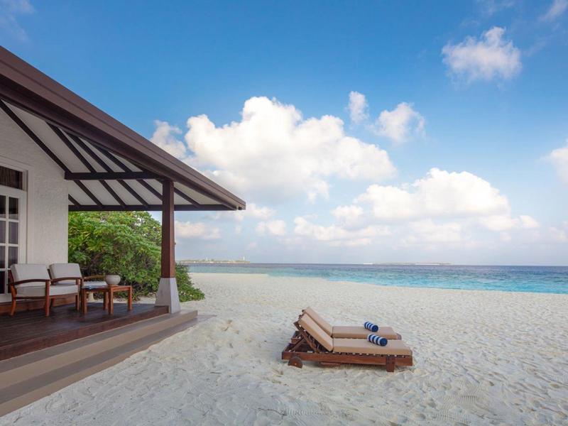 Villa de plage avec chaises longues sur sable blanc face à la mer bleue sous un ciel bleu.