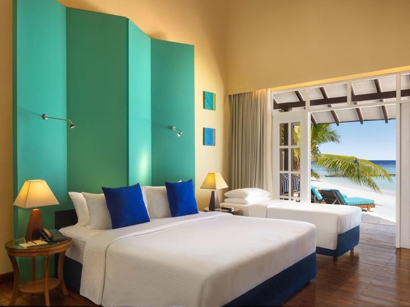 Chambre d'hôtel lumineuse avec deux lits, accents bleus et vue ouverte sur la plage.