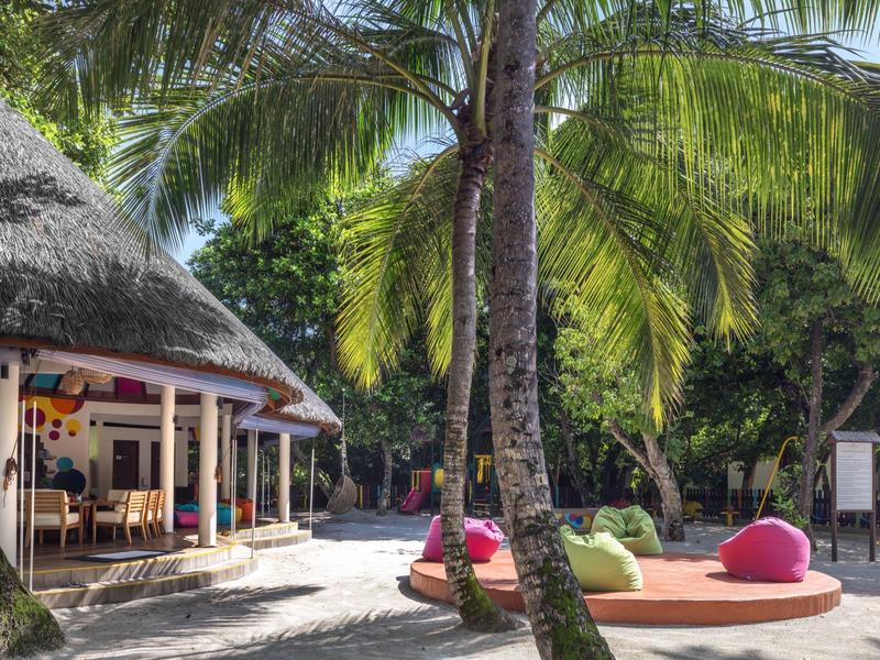 Bar de plage avec zone ombragée de palmiers, coussins colorés et ambiance détendue.