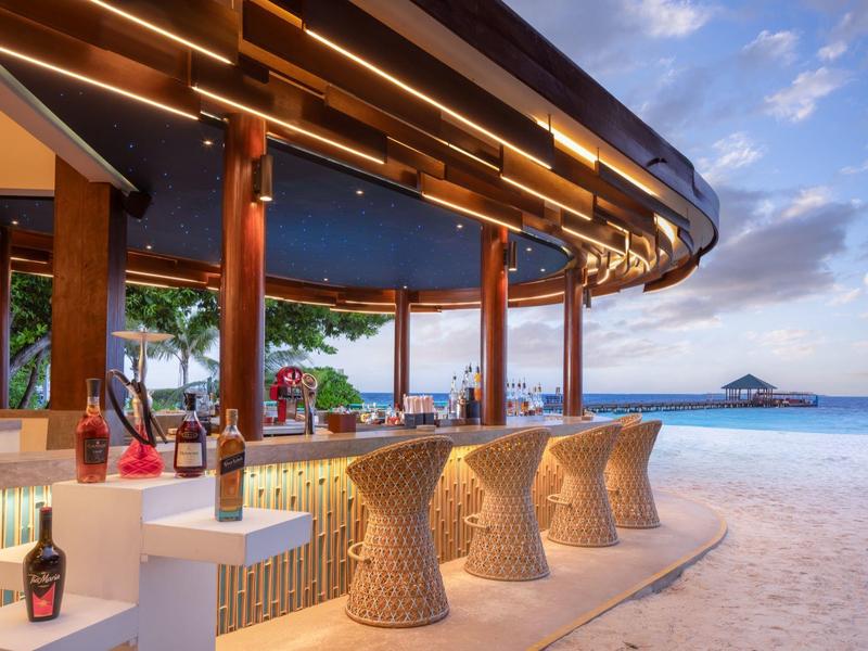 Bar de plage avec des tabourets en osier face à la mer, eau calme et ciel clair.