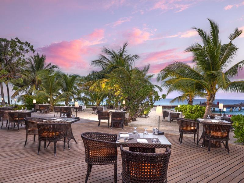 Restaurant en plein air avec chaises et tables sur une terrasse en bois avec vue sur la mer au coucher du soleil.