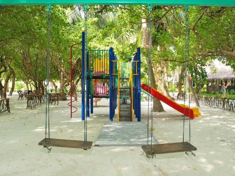 Kinderspielplatz mit buntem Klettergerüst, zwei Holzschaukeln und umgeben von Bäumen und Schatten.