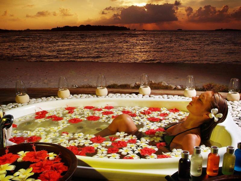 Frau entspannt in einem Blütenbad in einem Außen-Whirlpool bei Sonnenuntergang am Meer.