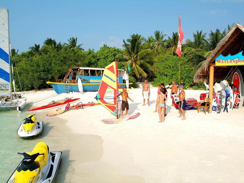 Strand mit weißem Sand, Segelboote, Jet-Skis und Menschen bei sonnigem Wetter und Palmen.