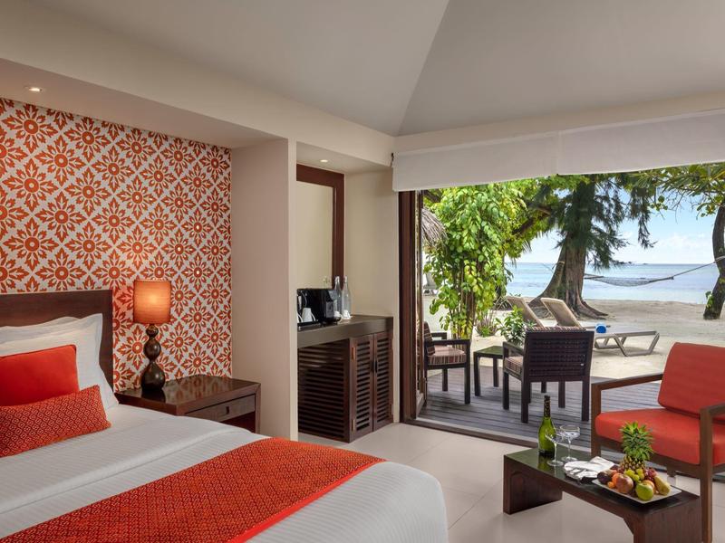 Gemütliches Zimmer mit weißem Bett, roten Akzenten, Balkon und Blick auf Palmen und Meer.